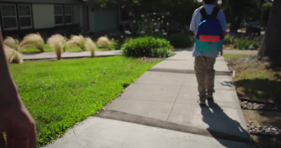 A mother sees her child off as he walks to school on the sidewalk at midday carrying a backpack