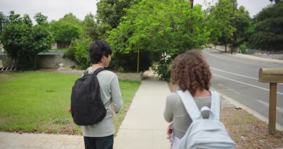 Two teenagers a boy and a girl are walking on the sidewalk at midday talking and carrying backpacks