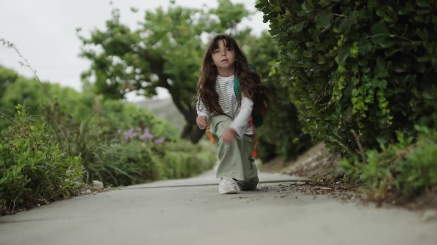 A young girl with long dark hair wearing a striped shirt and cargo pants kneels on a paved path tying her shoes then stands and runs away
