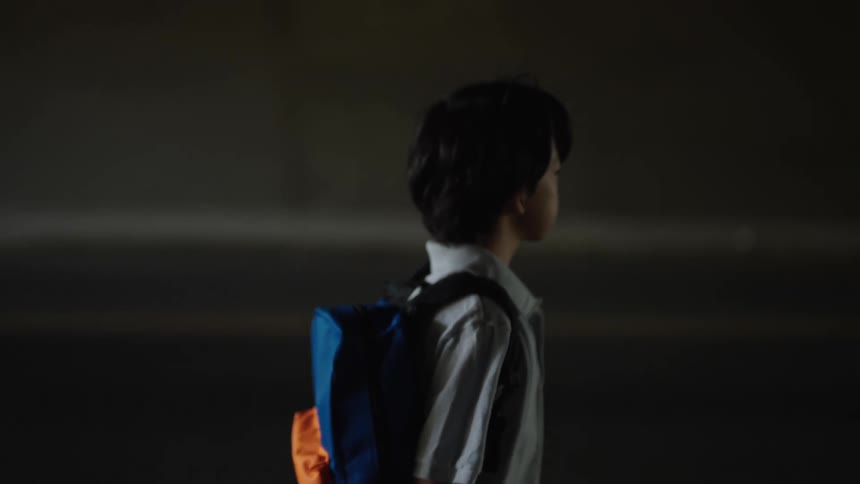 Young assian boy wearing a grey light weight jacket and a blue and orange colored back pack young boy is walking under what seems to be like an over pass or tunnel a red car zips by on the road next to where the boy is walking