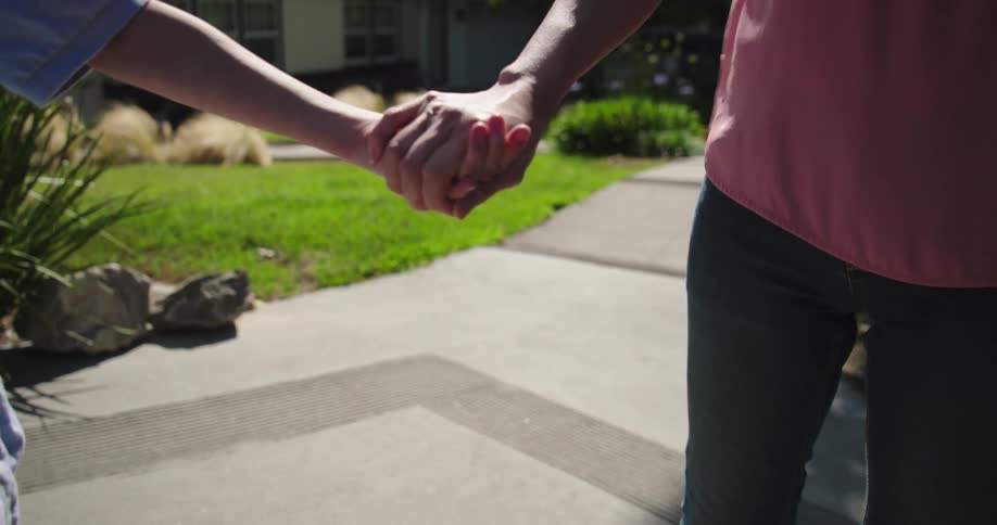A child and an adult walk down a sidewalk holding hands with a suburban house and yard in the background