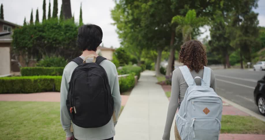 Two people with backpacks walk along a cracked sidewalk beside tangled branches and overgrown grass