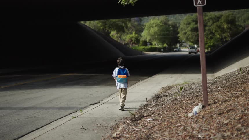 Boy wearing a backpack walks along a sidewalk passing under a bridge on a summer day