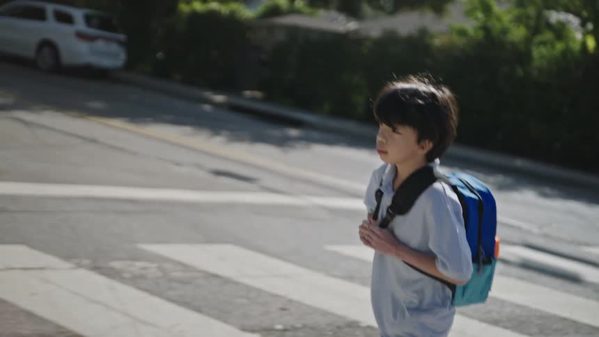 The video shows a child with a blue and orange backpack wearing a light blue polo shirt and khaki pants walking across a sunlit crosswalk they look left and right before proceeding with cars visible in the background