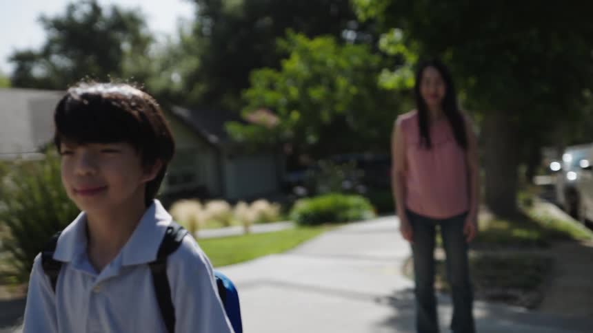 The video shows a child with a blue and orange backpack and a woman walking along a sunlit street with houses and trees in the background the child walks ahead while the woman watches