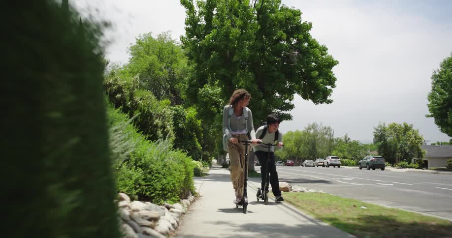 Two teenagers a boy and a girl are riding scooters on the sidewalk at midday each carrying a backpack