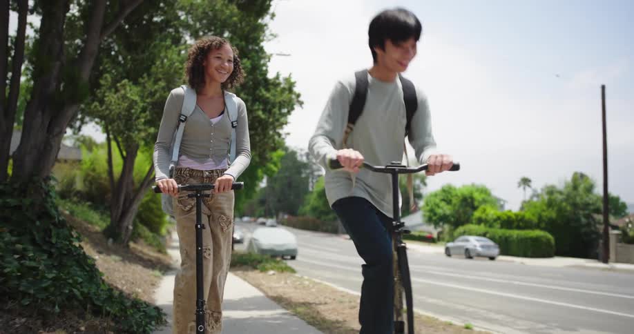 Two young individuals both wearing backpacks are riding scooters along a sidewalk next to a road the person in the foreground has curly hair and is wearing a light grey top and cargo pants while the person behind them has dark straight hair