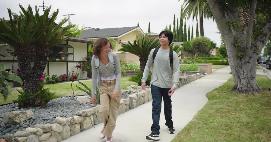Two young people with backpacks walk and run along a concrete sidewalk past a suburban house with a well maintained lawn and blooming pink flowers