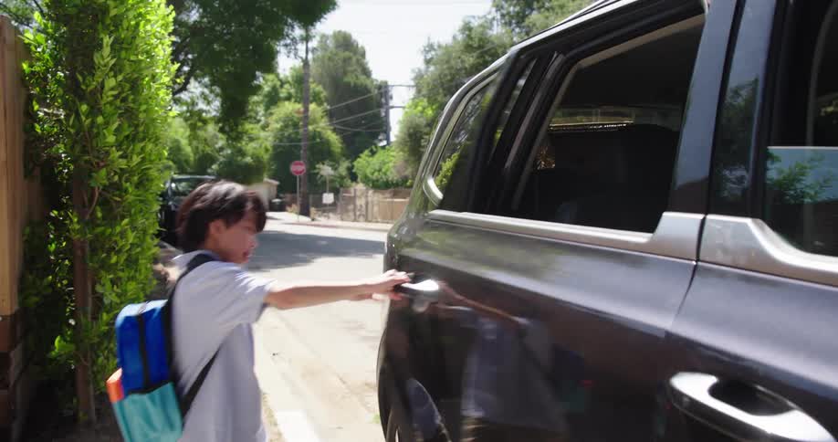 A child with a blue and green backpack runs out of a house opens the rear door of a black suv and the vehicle pulls away on a tree lined residential street
