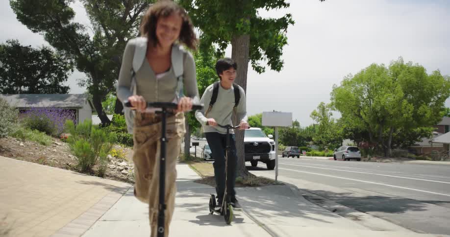 Two teenagers riding scooters with happy expressions on a suburban sidewalk
