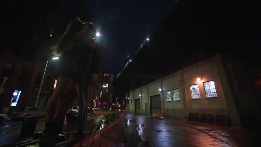 The video shows a couple embracing and standing close to each other on a wet street at night the scene is illuminated by the distant city lights and streetlights creating reflections on the wet ground