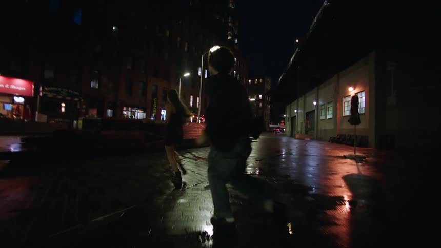 The video shows a couple walking at night on a wet street illuminated by streetlights and the glow from nearby buildings they briefly turn to face each other talking and laughing before continuing their walk