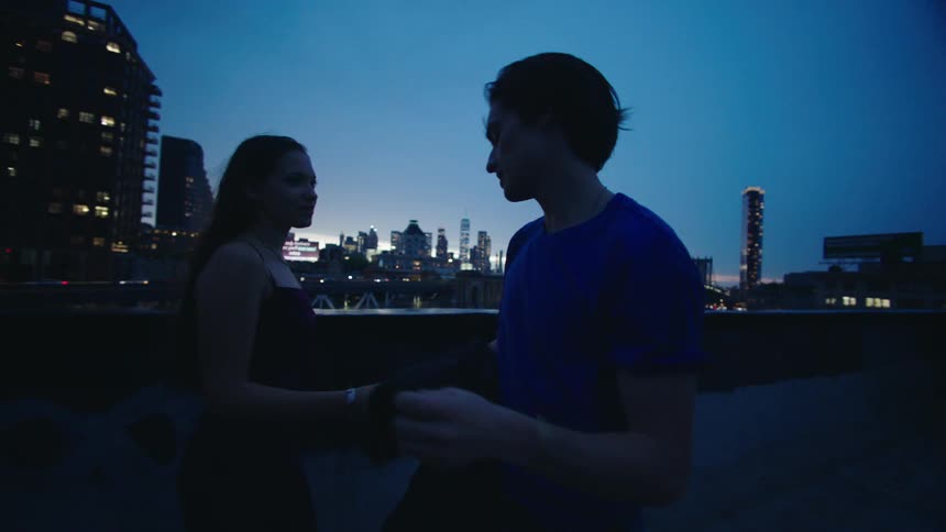 Man wraps up his girlfriend as they stand on a terrace with a panoramic view of a city at night