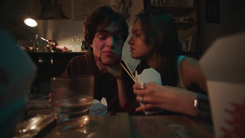 The video shows a closeup of a young couple eating a meal together in a dimly lit indoor setting they appear to be sharing food from a takeout container with chopsticks and the woman playfully feeds the man