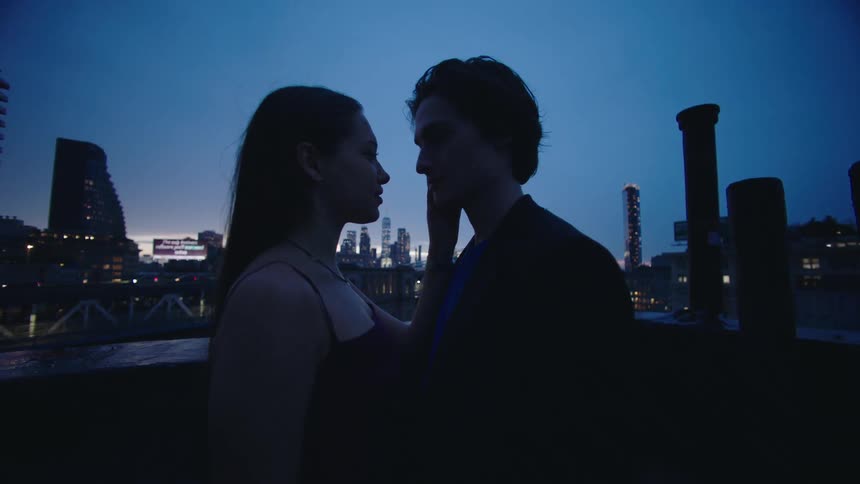 A man and a woman are passionately kissing on a rooftop with the city view in the background in the evening