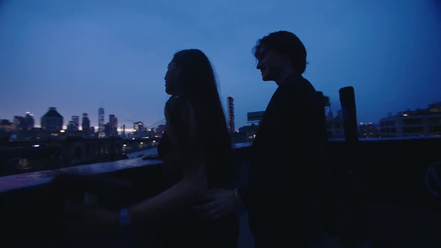 A couple runs handinhand on a rooftop at dusk with a city skyline and bridge in the background