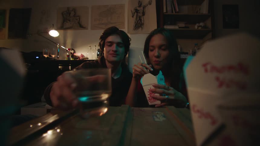 A young couple shares a meal from chinese takeout containers at a table with drawings on the wall behind them