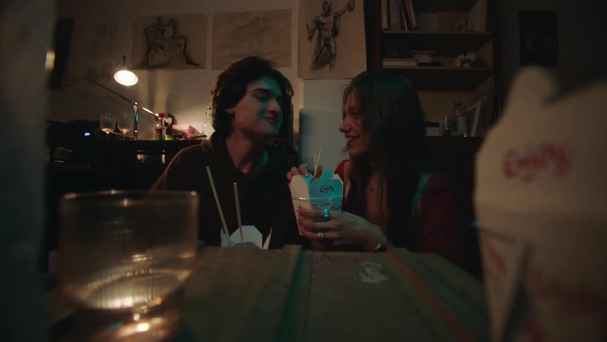 A young couple shares a meal from chinese takeout containers in a dimly lit room with drawings on the wall behind them