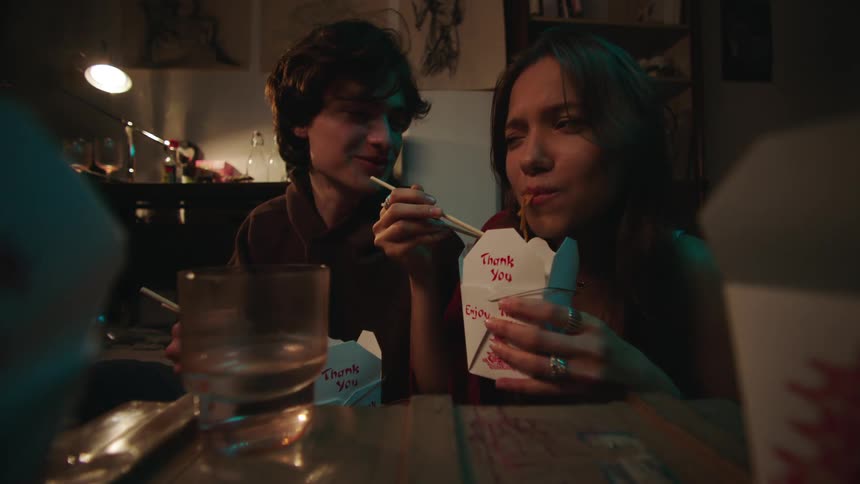 Two young adults a male and a female are seated at a table in a dimly lit room enjoying a meal from takeout containers the female uses chopsticks to eat from a white box labeled thank you enjoy