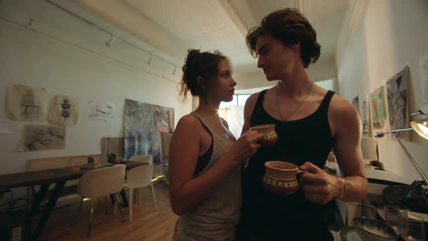A couple shares an intimate moment in a welllit spacious apartment filled with art the woman wearing a tank top and patterned shorts wraps her arms around the man who is holding a mug and gently kisses her forehead