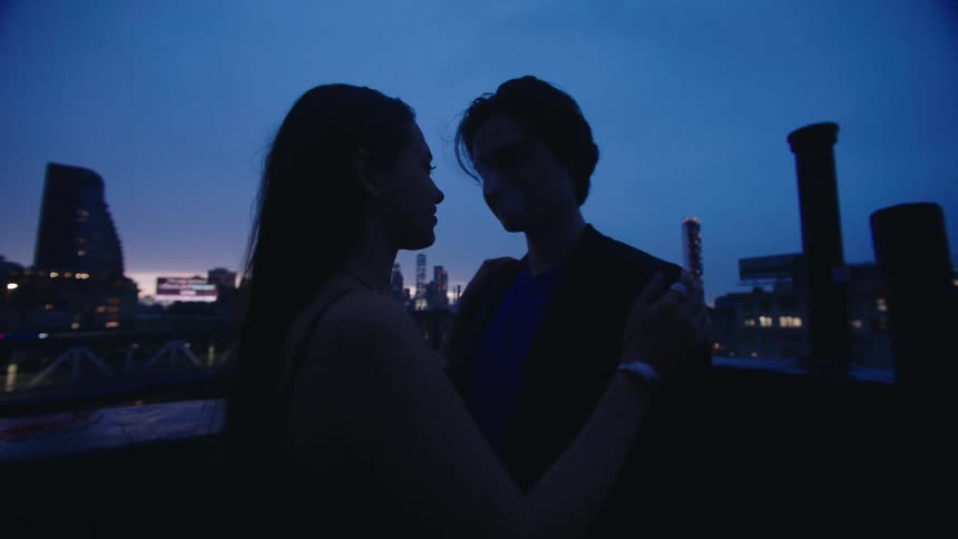 Couple standing on a roof with a view of a suspension bridge and the city skyline move closer for a kiss at night