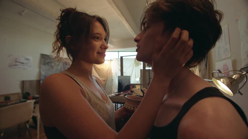 The video shows a tender moment between a couple in what appears to be an art studio the woman is wearing a lightcolored top and patterned bottoms and she caresses the mans face and hair while holding a small decorative cup