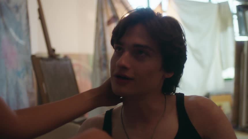A woman caresses a young mans face in a dim cluttered room their reflection visible in a mirror at the back