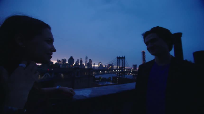 A couple stands on a rooftop at dusk silhouetted against a city skyline and a bridge