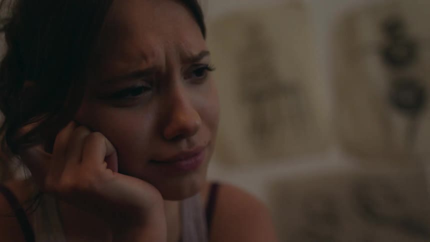 A young woman with dark hair sits in a dim room speaking gently to someone off screen with drawings pinned to the wall behind her
