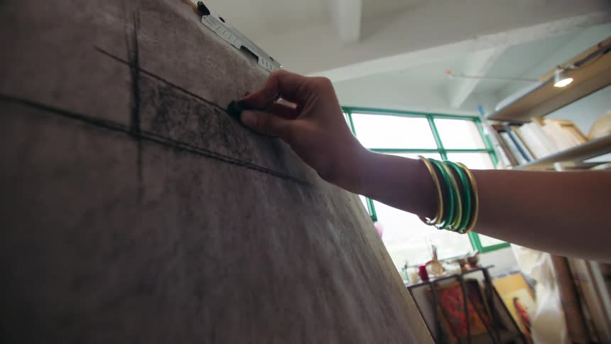 A hand with green and gold bangles drawing with charcoal on a large piece of paper mounted on an easel