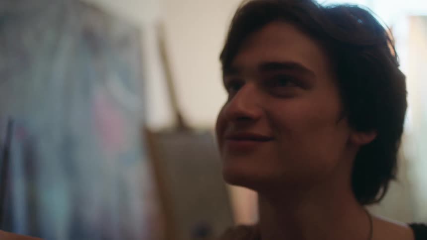 A young man with dark hair and a focused expression looks upward while painting with an easel and canvas behind him