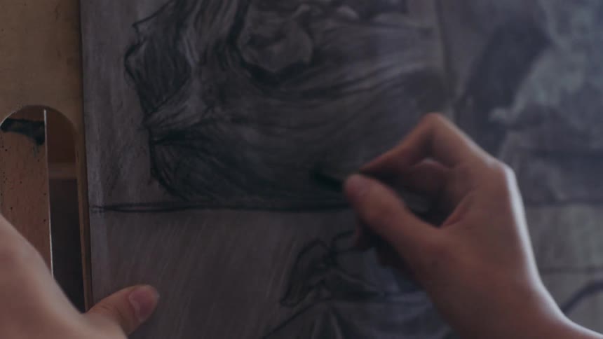 A persons hands are actively drawing a charcoal portrait on a canvas with multiple bracelets on one wrist