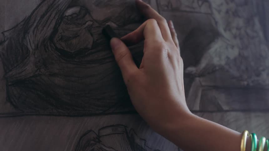 A hand wearing gold bangles shades a sketch with a small stick of drawing charcoal