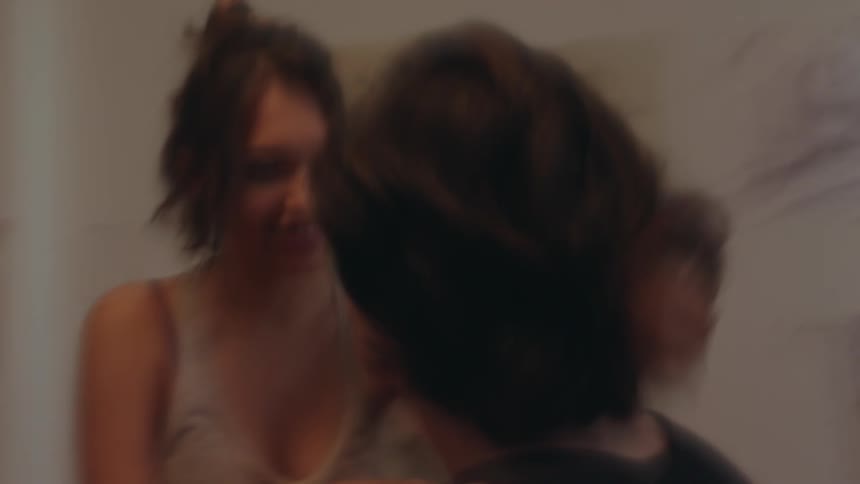 Two individuals are shown in an indoor setting one person with dark hair pulled back is wearing a dark top and the other with lighter hair is wearing a lightcolored top with a dark bra strap visible they appear to be in close proximity