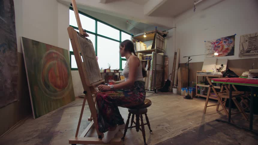 The video shows a woman with dark hair wearing a white tank top and patterned pants sitting on a stool and drawing on an easel in an art studio she takes a break to drink from a glass then stands up to continue her work