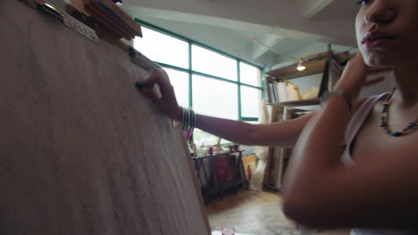 A young woman sets up a sheet of paper on an easel in a sunlit painting studio