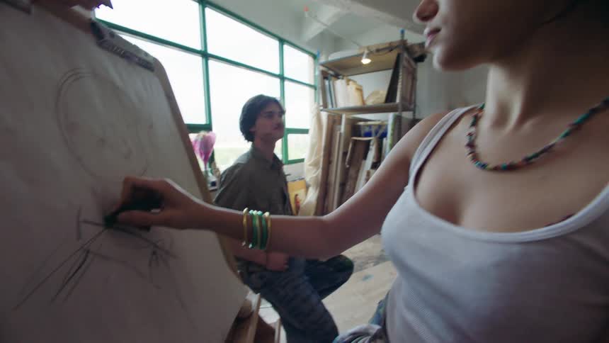 A young woman draws on an easel in an art studio while another woman watches