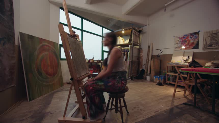 An artist sits on a stool painting at an easel in a cluttered studio filled with brushes canvases and finished artwork