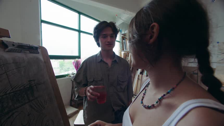 A person paints on a large canvas in a welllit studio as a man enters and gently hands her a beverage