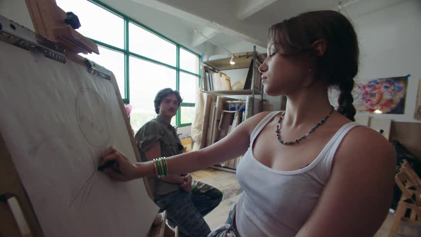A person sketches on a large sheet of paper at an easel in a sunlit art studio with large windows and shelves stocked with supplies while another person observes nearby