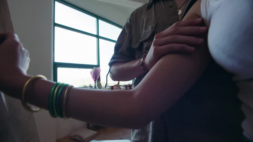 The video shows a woman wearing bracelets drawing on a canvas with a man standing close by the man gently touches the womans face and neck as they share an intimate moment in what appears to be an art studio with large windows