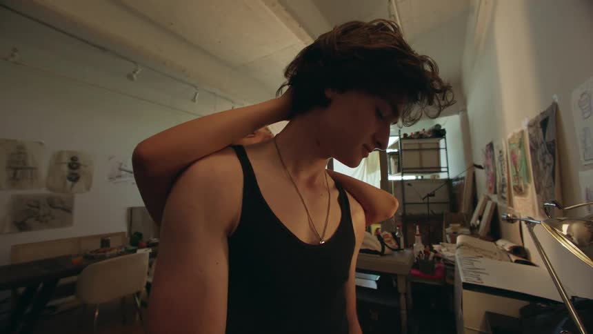 A young man in a tank top stands in an art studio while a female gently wraps her arms around his neck from behind