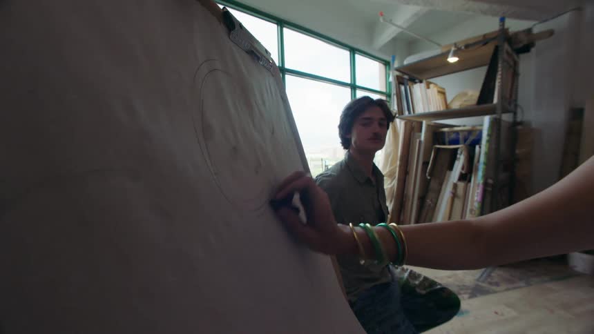 A woman is sketching a man posing with charcoal