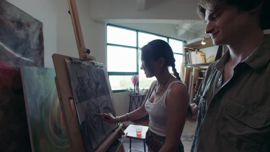 A woman concentrates while drawing on a large canvas in a sunlit art studio as a man walks up to her