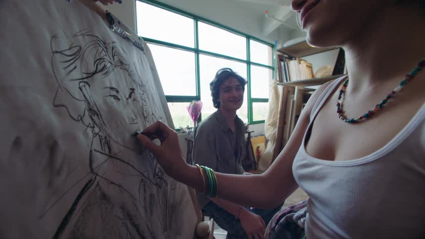 A young woman with braided hair and stacked bracelets is drawing a portrait on an easel while a young man watches her from behind