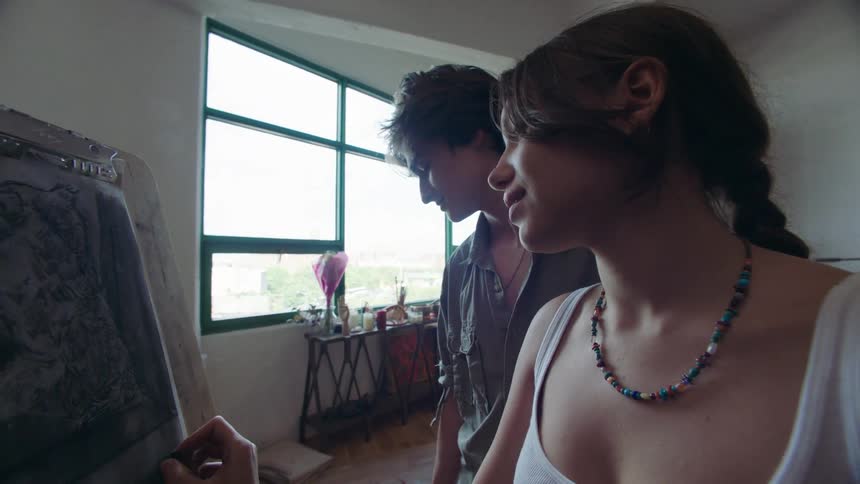 A woman paints on paper using charcoal at an easel while a man enters the room hands her a beverage and kisses her on the lips