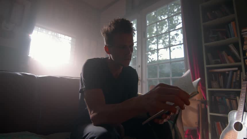 A young man is in a dark room and is turning the pages of a small book while sitting on a sofa