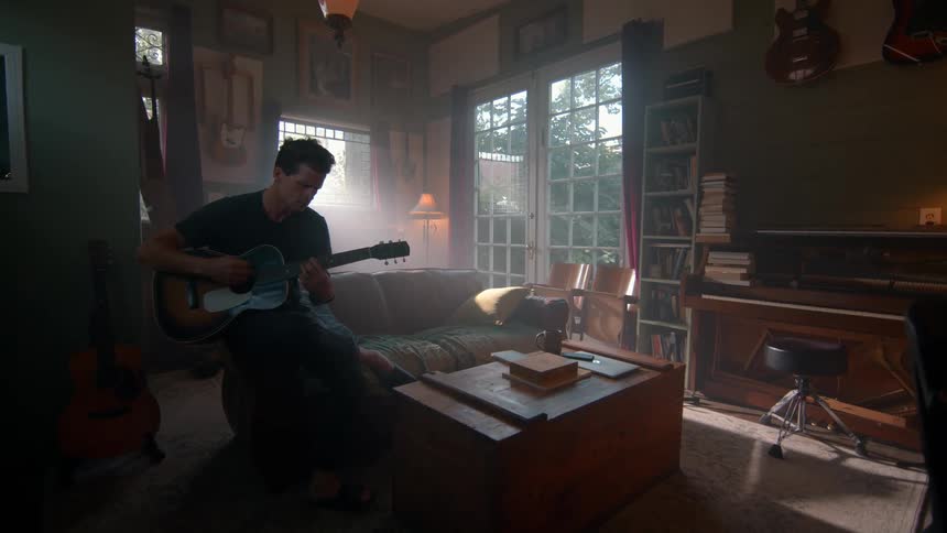A person is playing an acoustic guitar in a cozy room filled with musical instruments books and a piano with sunlight streaming through large windows