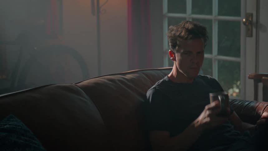 A person drinks from a mug while seated on a brown leather couch in a dim room with a bicycle by the wall and a reflective glass door