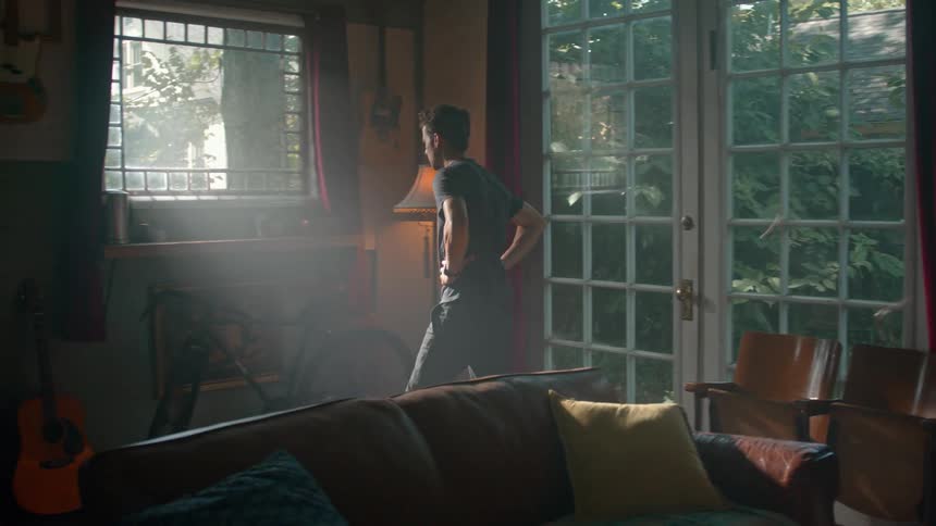 Sunlight streams through a window onto a person standing in a room filled with guitars a bicycle and a sofa